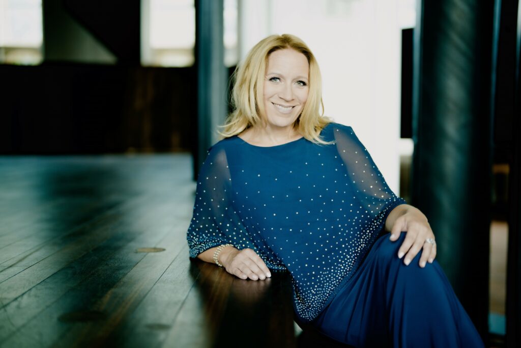 A blonde-haired woman in a blue dress smiling at the camera. 